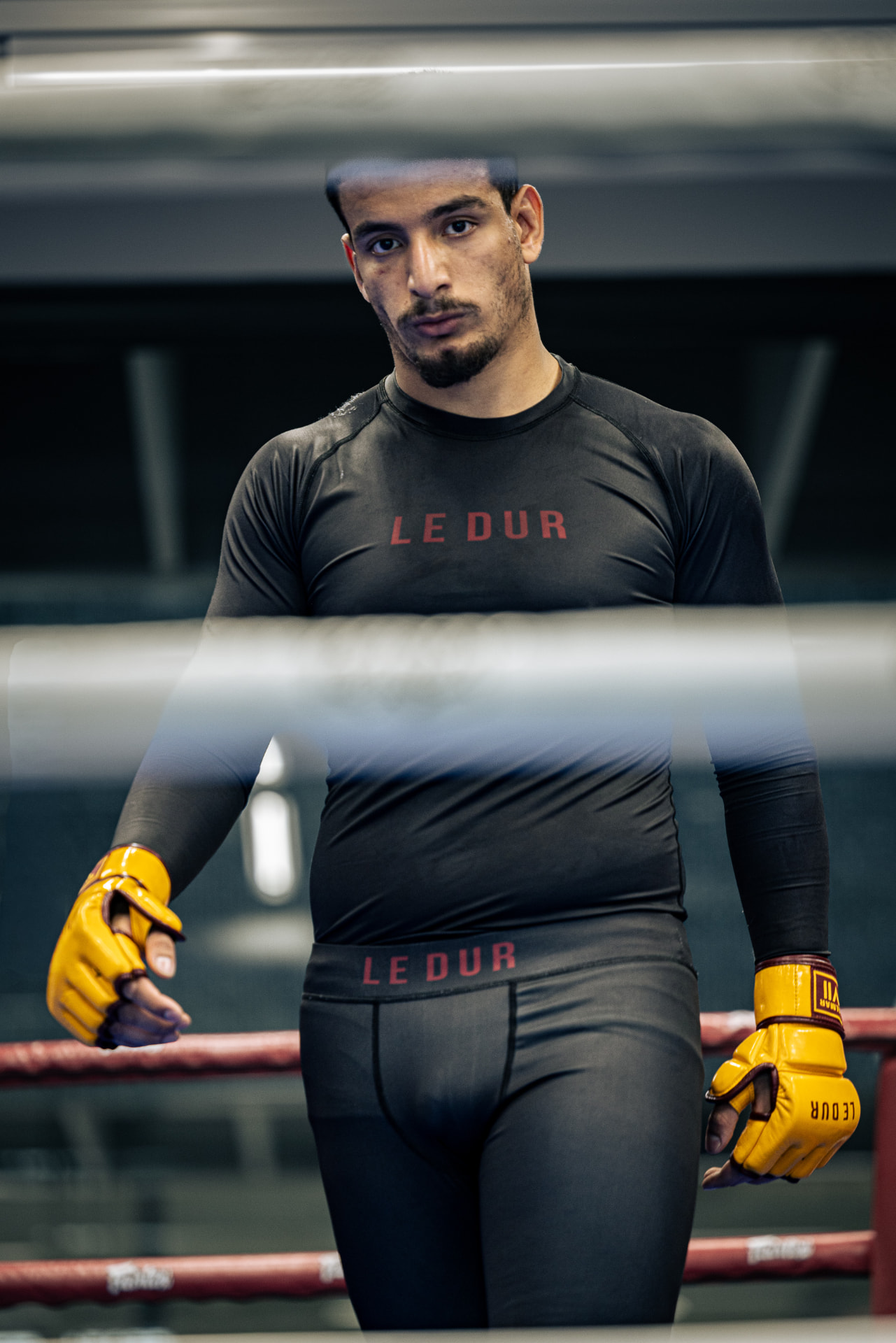 Murtada Khlaifawi standing in a boxing ring behind the ropes, wearing a black LE DUR rash guard, black LE DUR compression shorts and yellow LE DUR MMA gloves.
